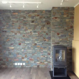 Ściana wykończona szarą mozaiką kamienną z elementami brązu i pomarańczy, z widocznymi gniazdkami elektrycznymi u dołu oraz nowoczesnym piecem wolnostojącym z czarną obudową i szybą, na drewnianej...