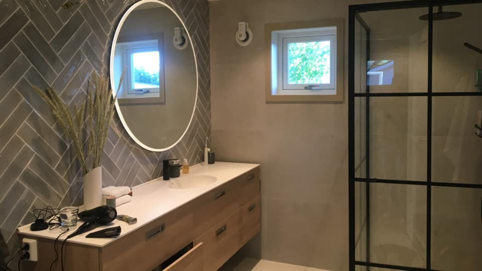 Nowoczesna łazienka z szarymi płytkami ułożonymi w jodełkę, okrągłym lustrem z podświetleniem LED, drewnianą szafką pod umywalką z otwartą szufladą i kabiną prysznicową z czarnymi szprosami.