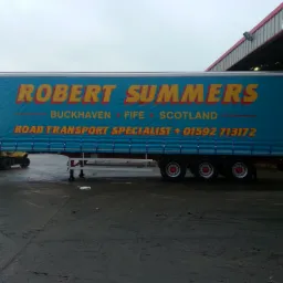 Niebieska naczepa z żółto-czerwonym nadrukiem nazwy firmy 'ROBERT SUMMERS' i danymi kontaktowymi, zaparkowana na mokrym placu przed halą.