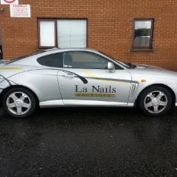 Srebrny samochód z grafiką i logo 'La Nails Boutique' zaparkowany na mokrym asfalcie w Dąbrowie Górniczej, na tle ceglanego budynku z oknami.