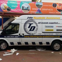 MPG Michał Pawelczyk - Okolicznościowe oklejenie folią reklamową białego samochodu dostawczego Ford Transit z logo firmy oponiarskiej, widoczne ślady po oklejaniu na podłodze, w tle baner reklamowy firmy zajmującej się...