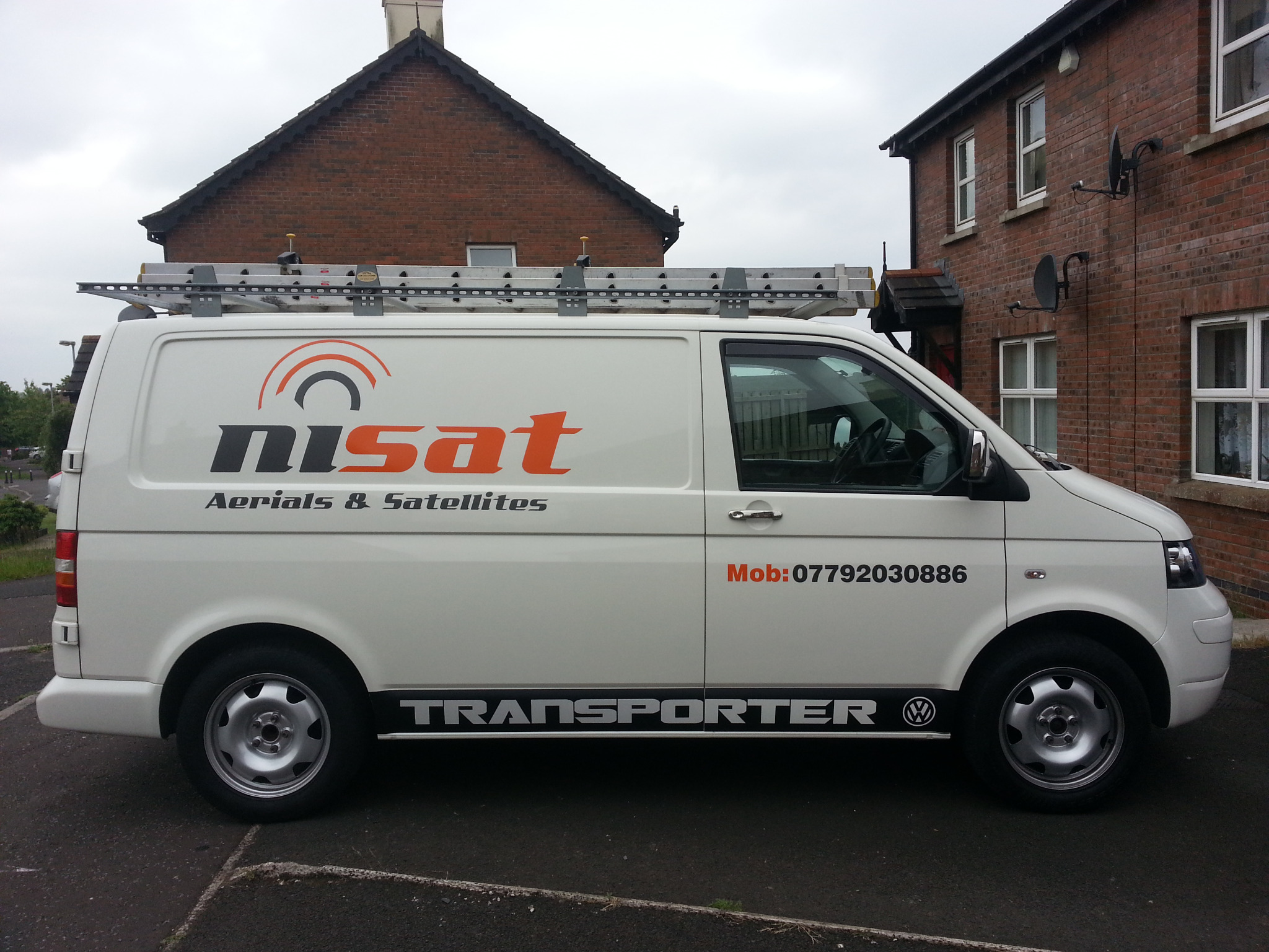 Biały van firmy instalującej anteny i satelity, z drabiną na dachu, zaparkowany przed domem z zamontowanymi antenami satelitarnymi.