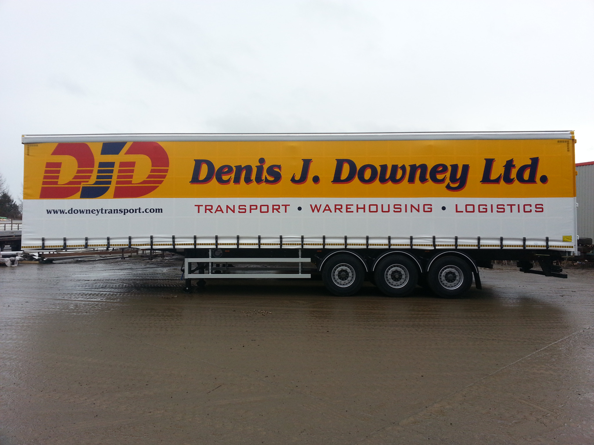 Żółta naczepa ciężarówki z logo i nazwą firmy Denis J. Downey Ltd., oferującej transport, magazynowanie i logistykę, zaparkowana na mokrym placu.