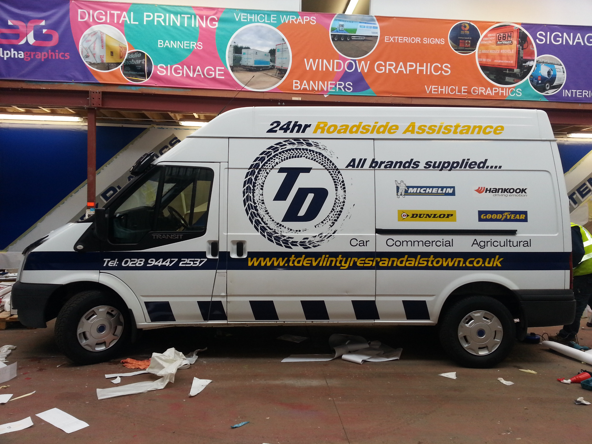 Okolicznościowe oklejenie folią reklamową białego samochodu dostawczego Ford Transit z logo firmy oponiarskiej, widoczne ślady po oklejaniu na podłodze, w tle baner reklamowy firmy zajmującej się...