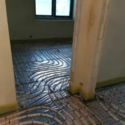 Montaż ogrzewania podłogowego z rurami grzewczymi ułożonymi na folii odblaskowej w surowym wnętrzu z widocznym oknem i fragmentem kolumny.