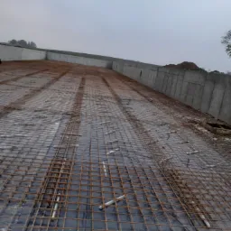 Siatka zbrojeniowa na czarnej folii, przygotowana pod wylewkę na fundamencie, widoczne betonowe ściany oporowe w tle, pochmurne niebo.