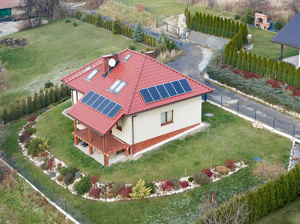 Dom jednorodzinny z czerwonym dachem i panelami słonecznymi, widok z góry, otoczony ogrodem z tujami i krzewami, z widocznymi oknami dachowymi i kominem.