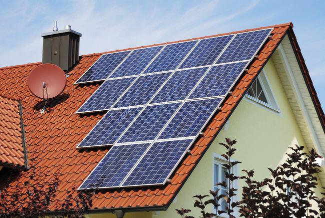 Dom z panelami słonecznymi na pomarańczowym dachu, z anteną satelitarną i kominem, na tle błękitnego nieba.