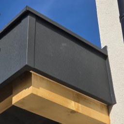 OLGIERD MORZYK REDE - Detal obróbki blacharskiej w kolorze grafitowym, zamontowanej na drewnianej konstrukcji przy ścianie budynku z widocznym orynnowaniem na tle błękitnego nieba.