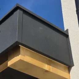 Detal obróbki blacharskiej w kolorze grafitowym, zamontowanej na drewnianej konstrukcji przy ścianie budynku z widocznym orynnowaniem na tle błękitnego nieba.