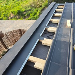 OLGIERD MORZYK REDE - Montaż obróbek blacharskich dachu z drewnianymi łatami w Krakowie. Widok z góry na fragment dachu z szarą blachą i elementami konstrukcyjnymi.