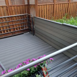 OLGIERD MORZYK REDE - Szary taras kompozytowy z metalową barierką i drewnianym ogrodzeniem. Widoczne kwiaty w donicach. Ujęcie z góry, podkreślające fakturę desek.