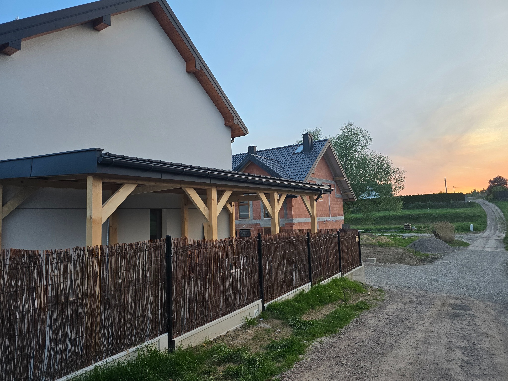 Nowoczesna pergola z drewnianą konstrukcją i trzcinową osłoną, przylegająca do elewacji domu. W tle widać budynek w trakcie budowy i drogę.