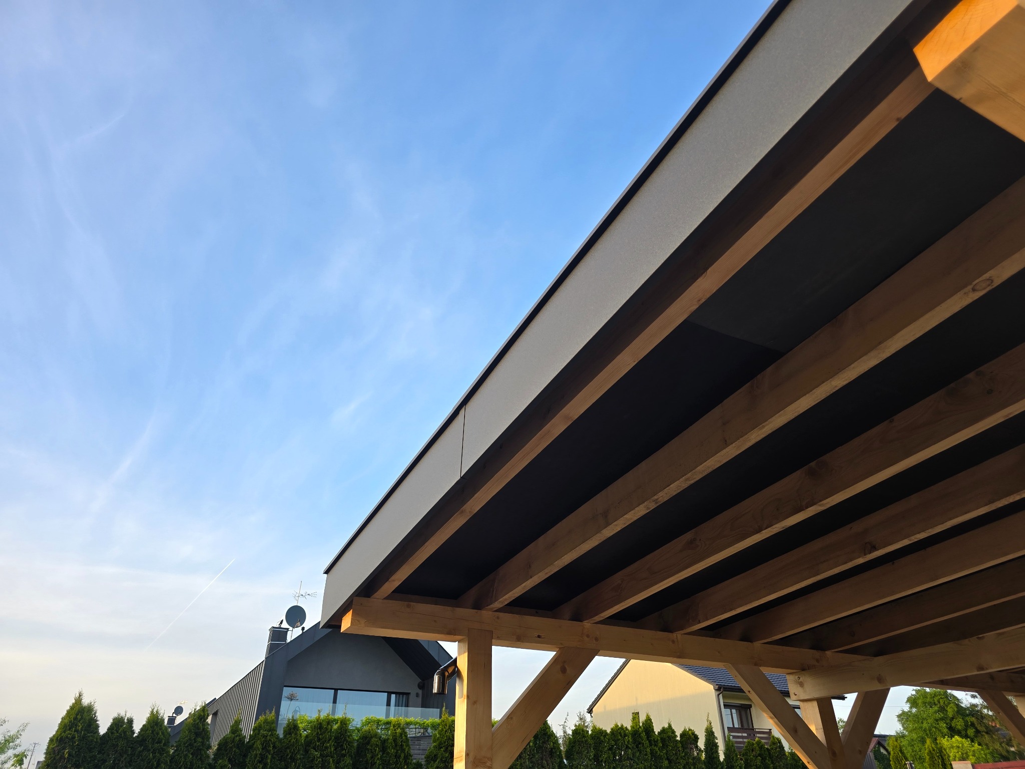 Drewniana konstrukcja z belek, prawdopodobnie zadaszenie lub pergola, widziana od dołu na tle błękitnego nieba. W tle widoczne domy i zieleń.
