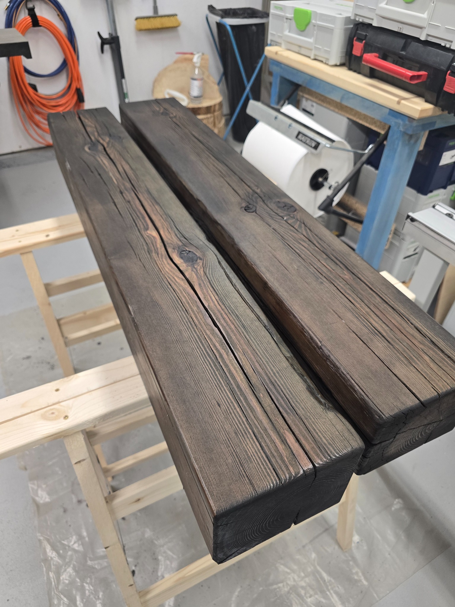 Dwa rustykalne, ciemnobrązowe belki z wyraźnym usłojeniem leżą na drewnianym koźle. Widoczne sęki i pęknięcia drewna. W tle narzędzia i akcesoria warsztatowe.