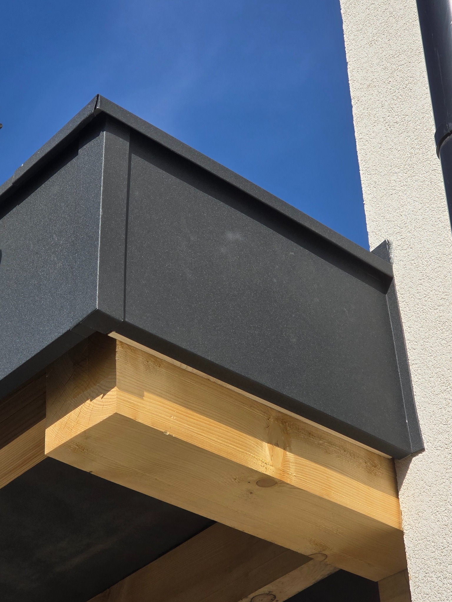 Detal obróbki blacharskiej w kolorze grafitowym, zamontowanej na drewnianej konstrukcji przy ścianie budynku z widocznym orynnowaniem na tle błękitnego nieba.