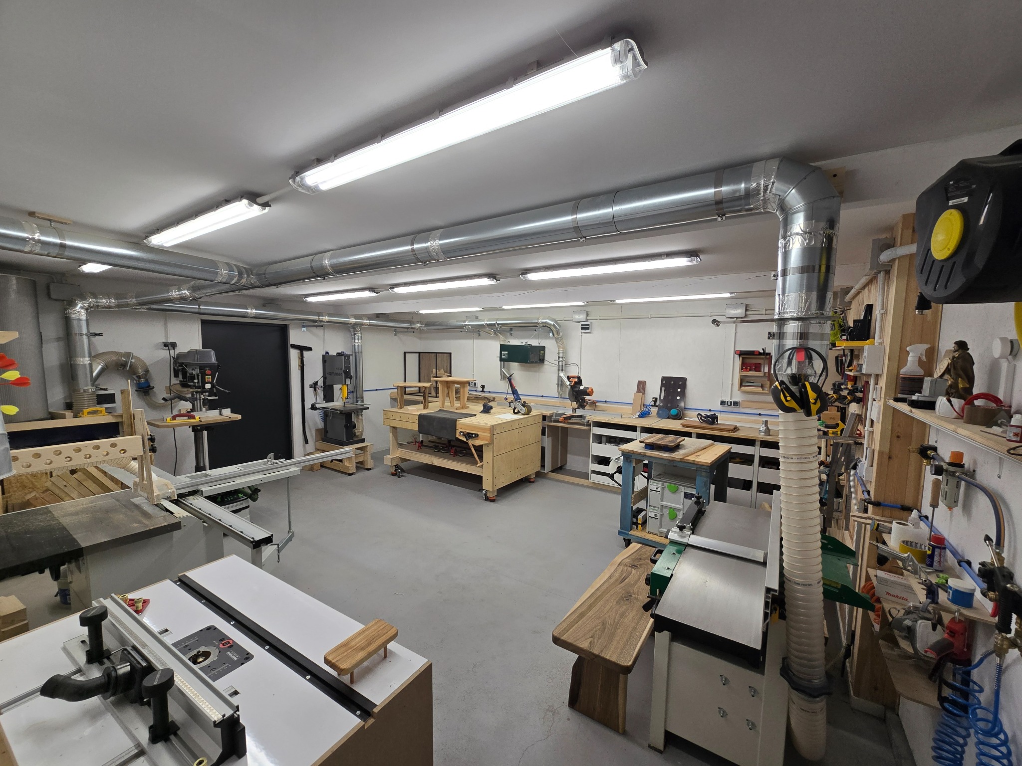 Przestronny warsztat stolarski z licznymi narzędziami i maszynami do obróbki drewna, oświetlony lampami jarzeniowymi i wyposażony w system wentylacji.