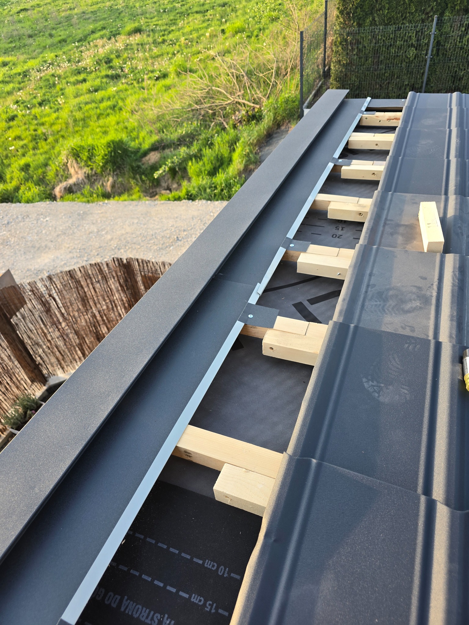 Montaż obróbek blacharskich dachu z drewnianymi łatami w Krakowie. Widok z góry na fragment dachu z szarą blachą i elementami konstrukcyjnymi.