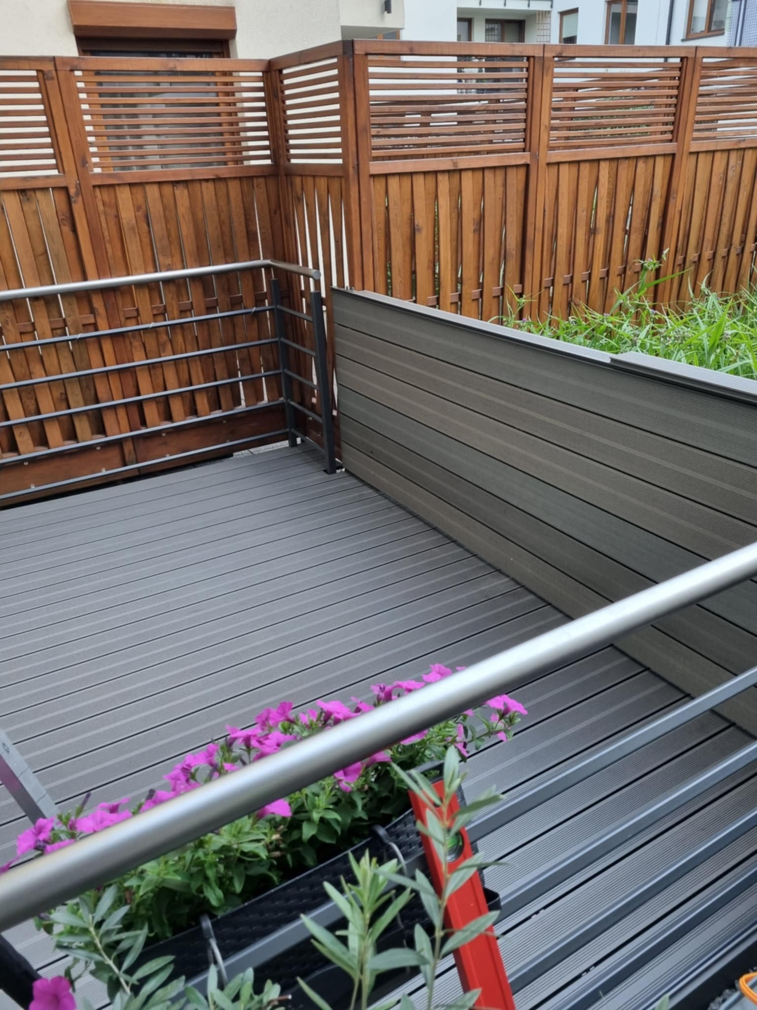 Szary taras kompozytowy z metalową barierką i drewnianym ogrodzeniem. Widoczne kwiaty w donicach. Ujęcie z góry, podkreślające fakturę desek.