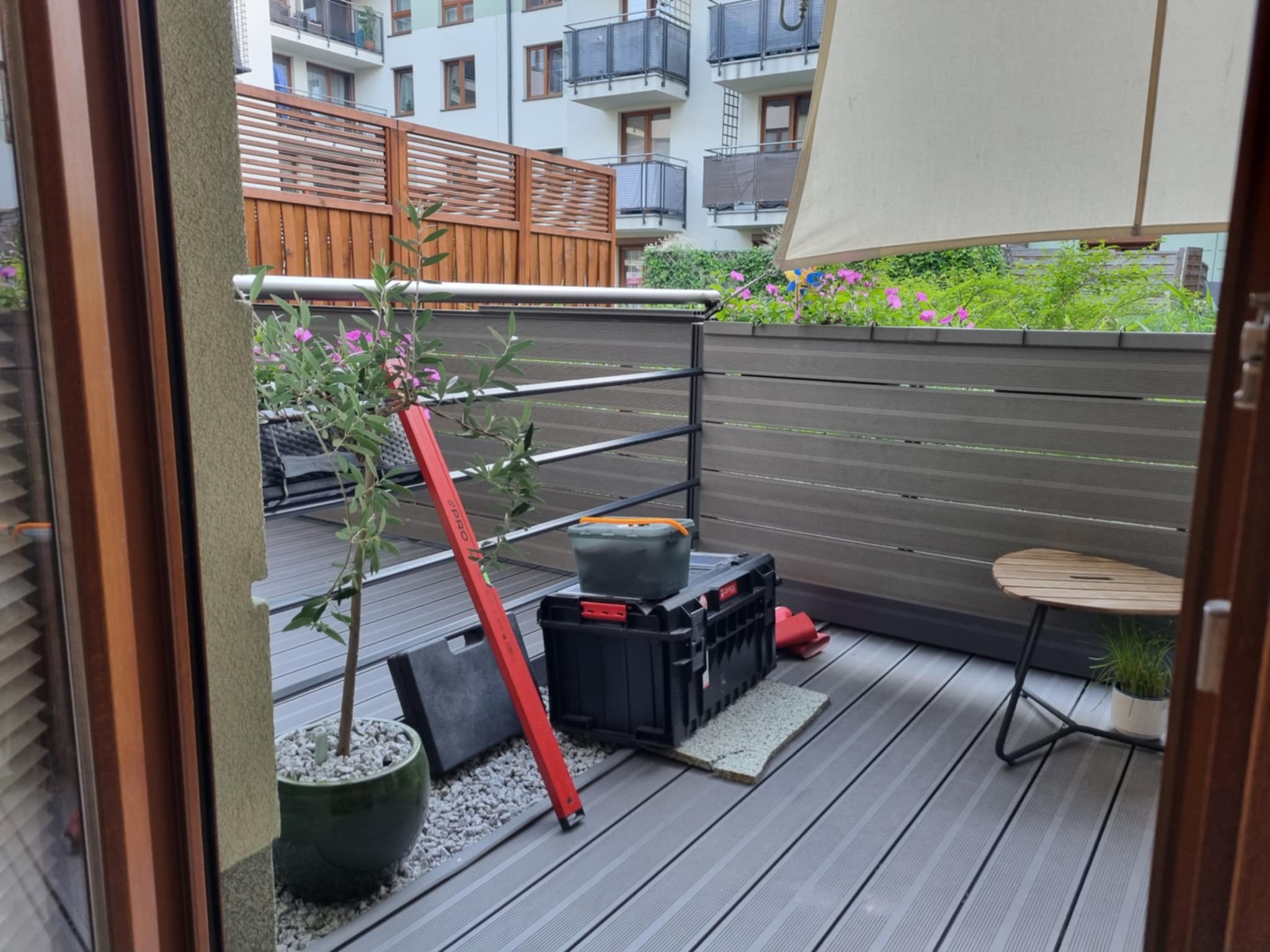 Nowoczesny balkon z szarą podłogą kompozytową, roślinami w donicach i skrzynką narzędziową. W tle widoczny budynek mieszkalny z drewnianymi elementami.