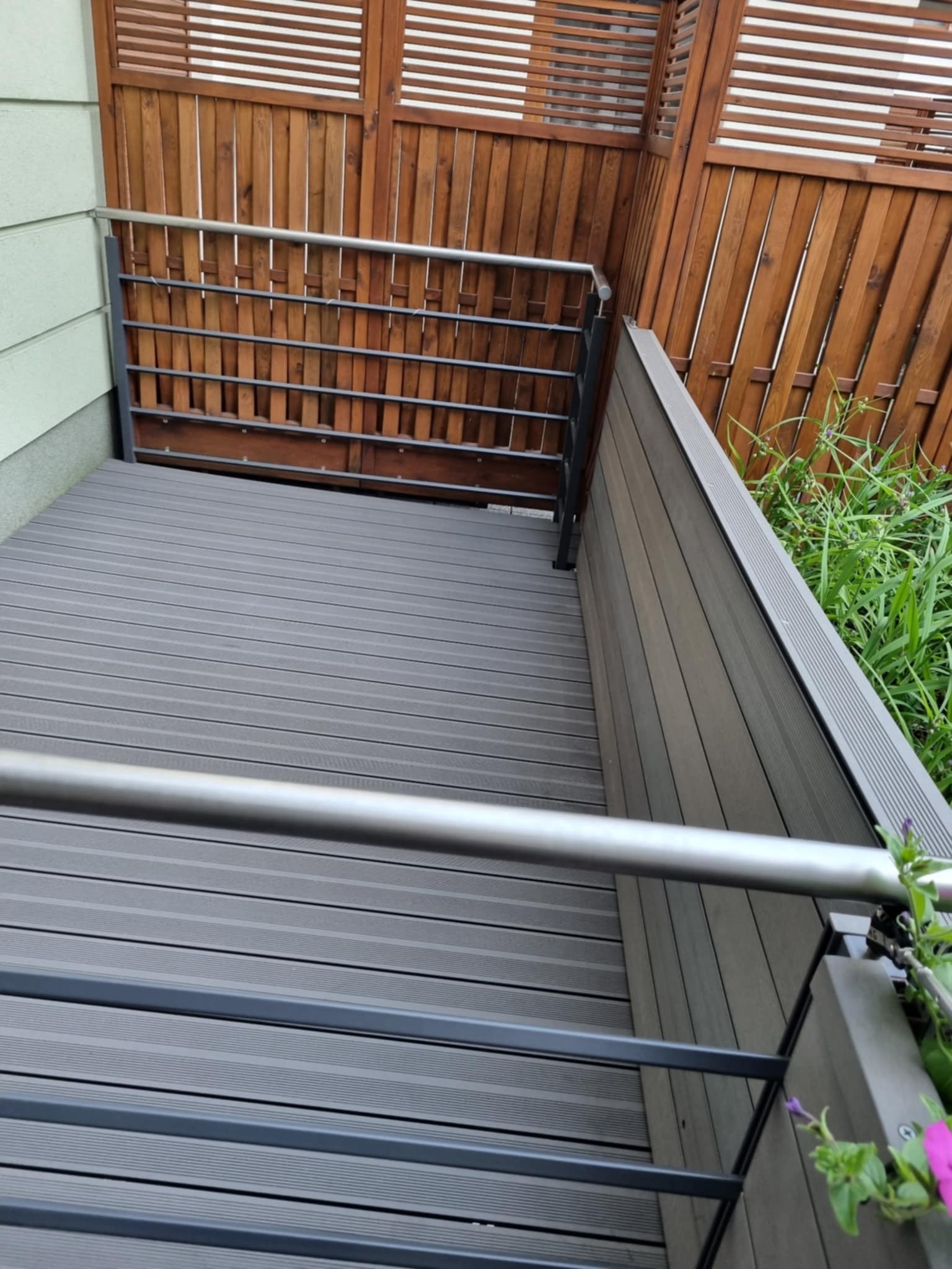 Szary taras kompozytowy z metalową balustradą i drewnianym ażurowym panelem, widok z góry, z elementami zieleni w tle. Nowoczesny design.