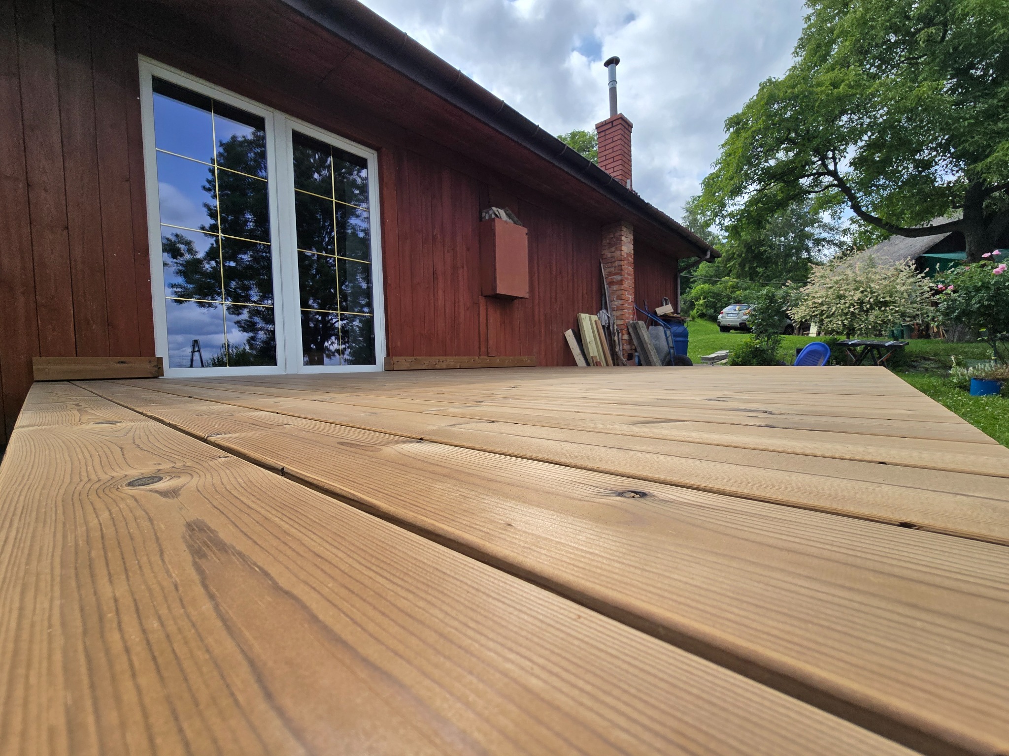 Nowy, drewniany taras przylegający do domu z brązową elewacją, widok z niskiej perspektywy podkreśla strukturę drewna i solidność wykonania. W tle zieleń ogrodu.
