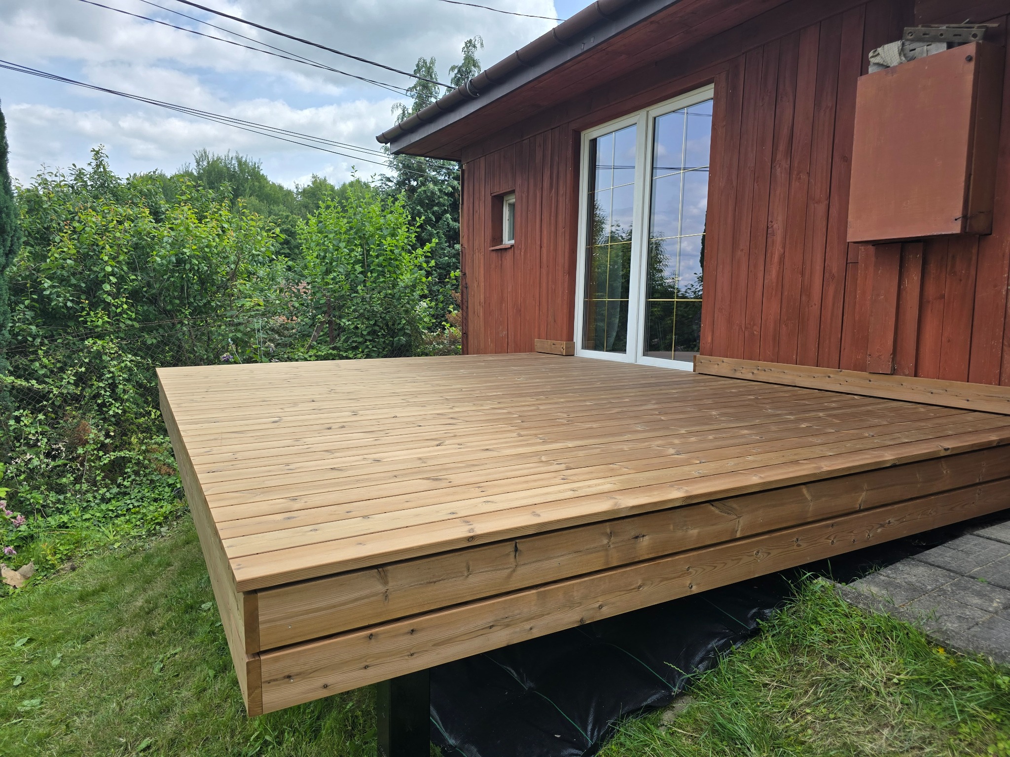 Jasny, drewniany taras przylegający do budynku z czerwonymi ścianami. Widoczne białe okna i zieleń w tle. Kąt ujęcia ukazuje solidną konstrukcję.