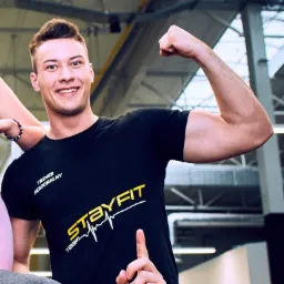 Uśmiechnięty mężczyzna w czarnej koszulce z napisem 'STAYFIT TEAM' prezentuje napięty biceps na tle siłowni.