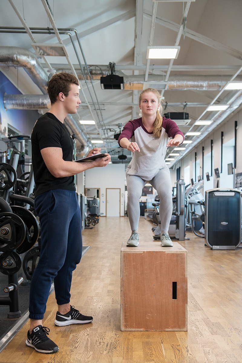 Kobieta wykonuje przysiad na skrzyni pod nadzorem trenera z tabletem w siłowni, widoczne maszyny do ćwiczeń w tle.