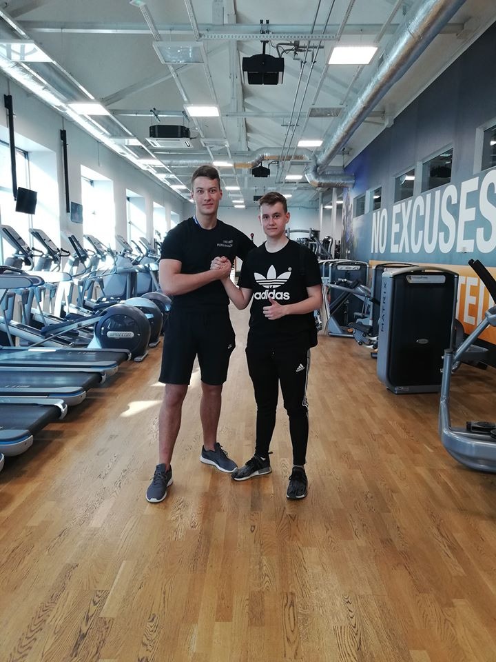 Trener personalny w czarnej koszulce i spodenkach w siłowni, ściska dłoń klienta po treningu, w tle rzędy bieżni i napis NO EXCUSES na ścianie.