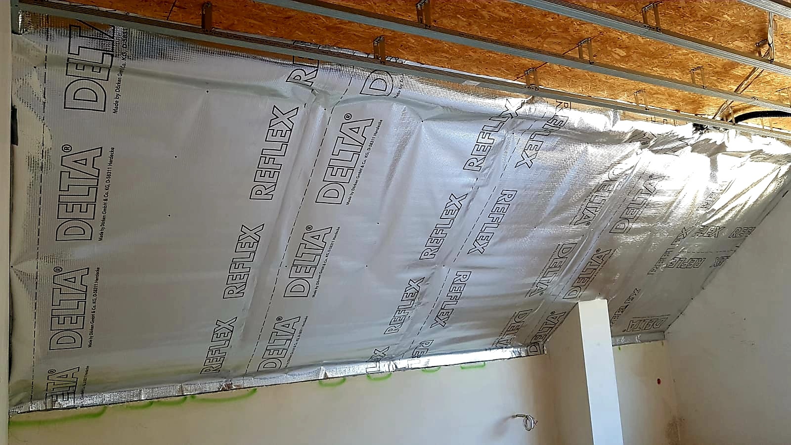 Montaż folii refleksyjnej Delta Reflex na poddaszu, widoczne metalowe profile konstrukcyjne pod sufitem i fragment ściany. Izolacja termiczna z nadrukowanym logo producenta.