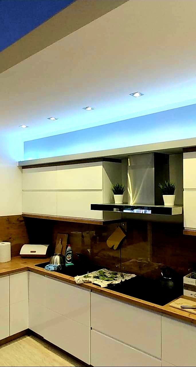 Nowoczesna kuchnia z białymi szafkami i drewnianymi blatami, oświetlona niebieskim światłem LED pod sufitem podwieszanym, z widocznymi punktowymi lampami sufitowymi.