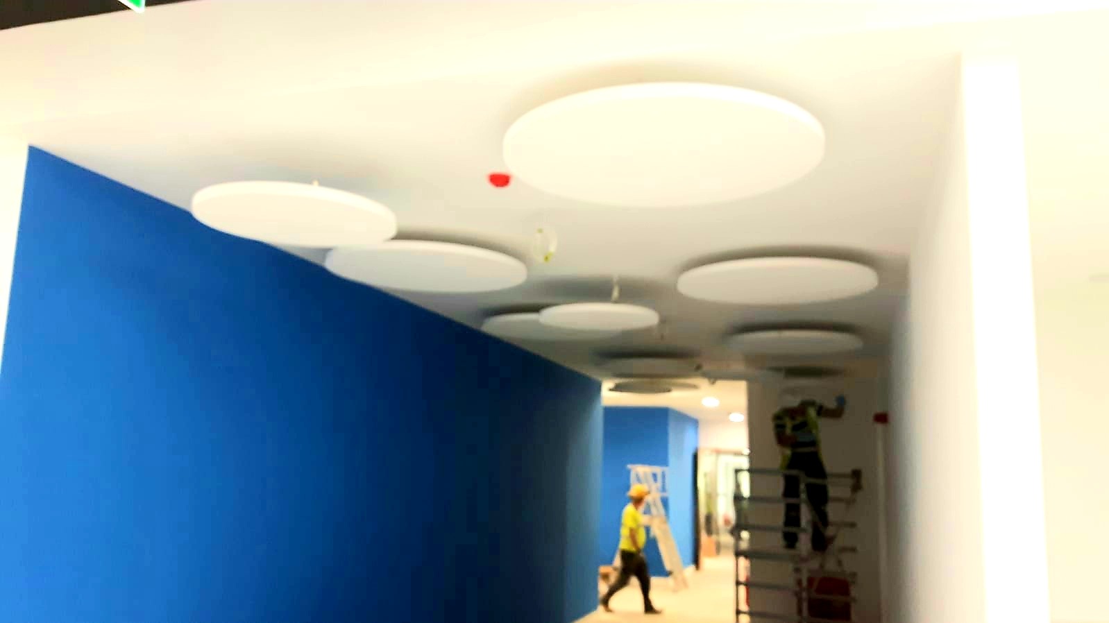 Instalacja okrągłych paneli akustycznych na suficie w biurze, widoczni pracownicy na rusztowaniach, niebieska ściana w tle.