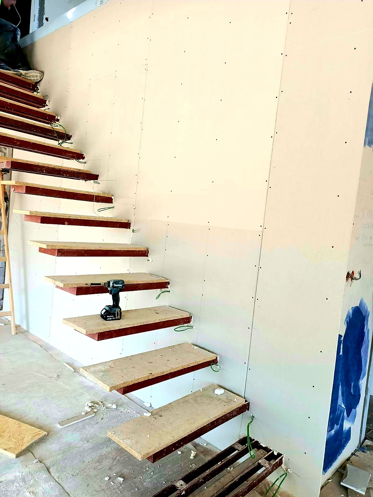 Konstrukcja schodów z płyt kartonowo-gipsowych na metalowej ramie, z widocznym wkrętakiem akumulatorowym Makita na jednym ze stopni, oraz fragmentem ściany pokrytej płytami g-k.