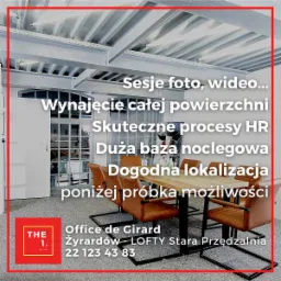 Jasne wnętrze biurowe z drewnianym stołem i skórzanymi krzesłami, surowy sufit z widocznymi instalacjami, okno z podziałami, w tle grafika na ścianie.