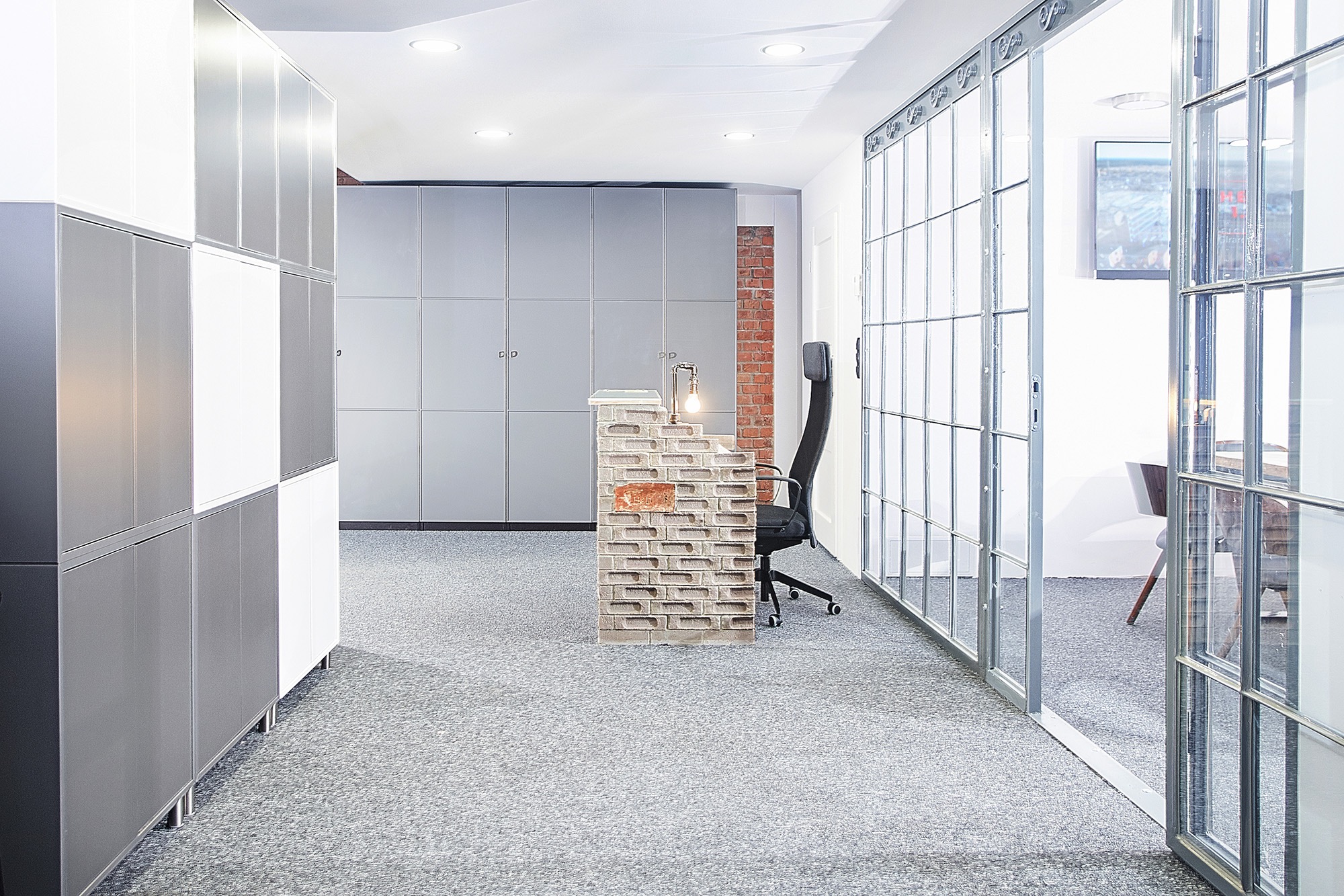 Nowoczesna recepcja w biurze z ceglaną ladą, oświetlona lampką biurkową, z widocznym krzesłem obrotowym i szafkami w tle, oddzielona szklaną ścianą z metalową ramą.