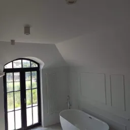 Nowoczesna łazienka na poddaszu z wolnostojącą wanną, grzejnikiem żeberkowym i dużym oknem z czarną ramą wychodzącym na zieleń.