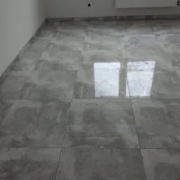 Ułożone szare płytki podłogowe imitujące beton z widocznymi fugami, fragment białej ściany z grzejnikiem i otworem w ścianie.