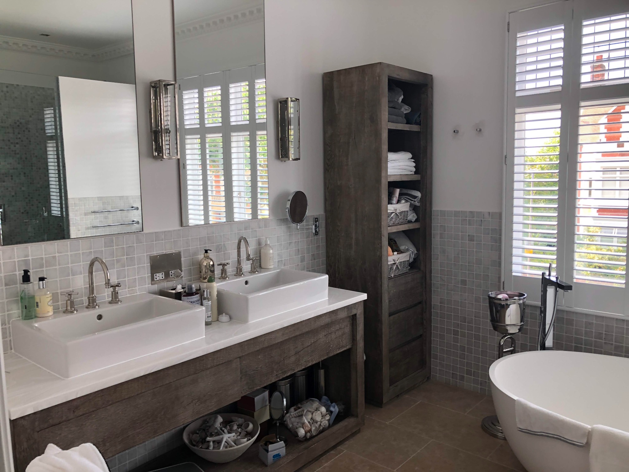 Elegancka łazienka z podwójną umywalką nablatową, drewnianą szafką z dekoracjami morskimi, kolumną z półkami na ręczniki oraz wolnostojącą wanną przy oknie z żaluzjami.