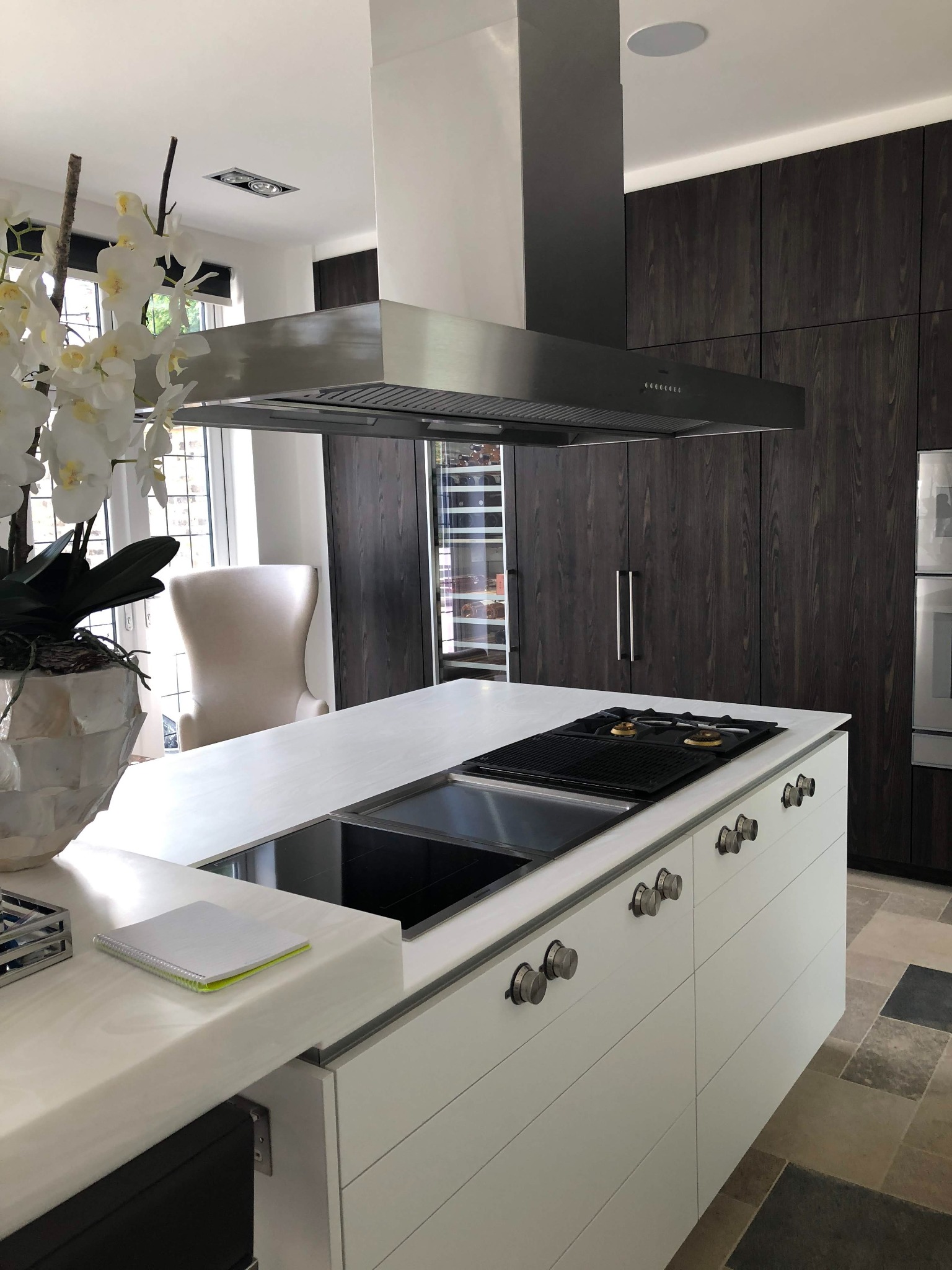 Nowoczesna kuchnia z wyspą, stalowym okapem i ciemnymi drewnianymi szafkami, na blacie leży notatnik z zielonymi elementami, podłoga wyłożona kamiennymi płytkami.