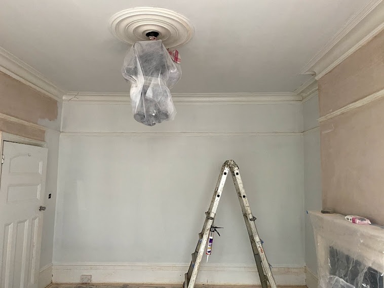 Pokój w trakcie odświeżania: ściany częściowo pomalowane na jasny odcień, zabezpieczony folią żyrandol pod ozdobnym plafonem, drabina aluminiowa z tubą silikonu, fragment ściany z widocznym gruntem.