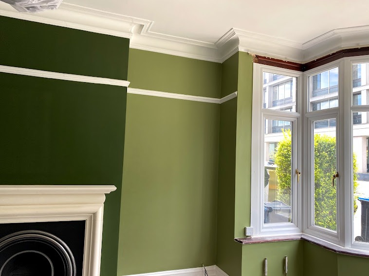 Wnętrze pokoju po odświeżeniu: ściany pomalowane na oliwkowy odcień zieleni, białe listwy przypodłogowe i sufitowe, widoczny fragment kominka z białym obramowaniem, okno wykuszowe z białymi ramami,...
