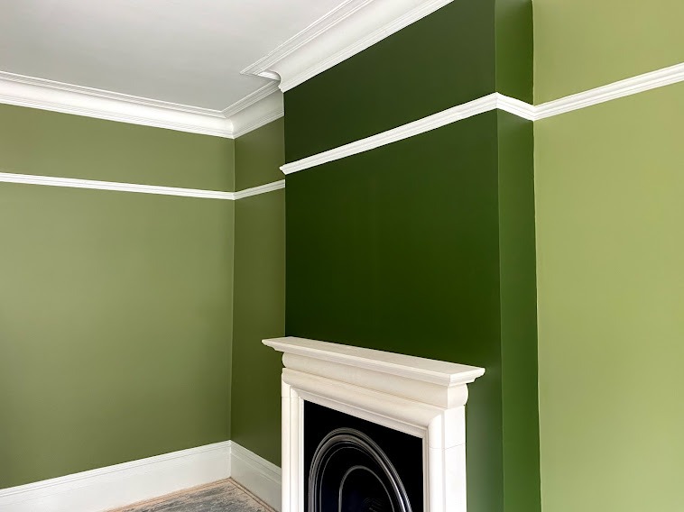Wnętrze po odświeżeniu: ściany pomalowane na dwa odcienie zieleni, ciemniejszy akcent przy kominku z białym obramowaniem, białe listwy przypodłogowe i sufitowe.