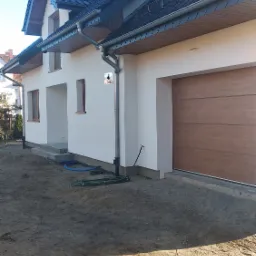 Świeżo otynkowany dom jednorodzinny z brązową bramą garażową i ciemnym dachem, widoczne niebieskie węże ogrodowe na piasku przed budynkiem.