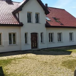 Dwukondygnacyjny dom jednorodzinny z jasną elewacją, czerwoną dach&oacute;wką i brukowanym podjazdem w słoneczny dzień.