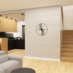 LA CASA STUDIO Iwona Wolska - Wnętrze domu z otwartą kuchnią z drewnianymi frontami, schodami i szarą sofą. Na ścianie zegar, a nad blatem wisząca roślina. Nowoczesny design.