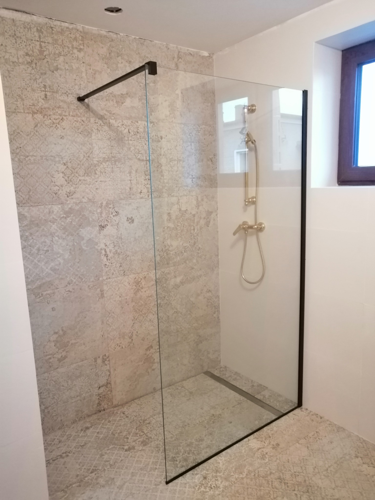 Nowoczesna kabina prysznicowa walk-in z czarnymi profilami i deszczownicą w odcieniach złota, zamontowana w wykończonej łazience z beżowymi płytkami o wzorzystej strukturze.