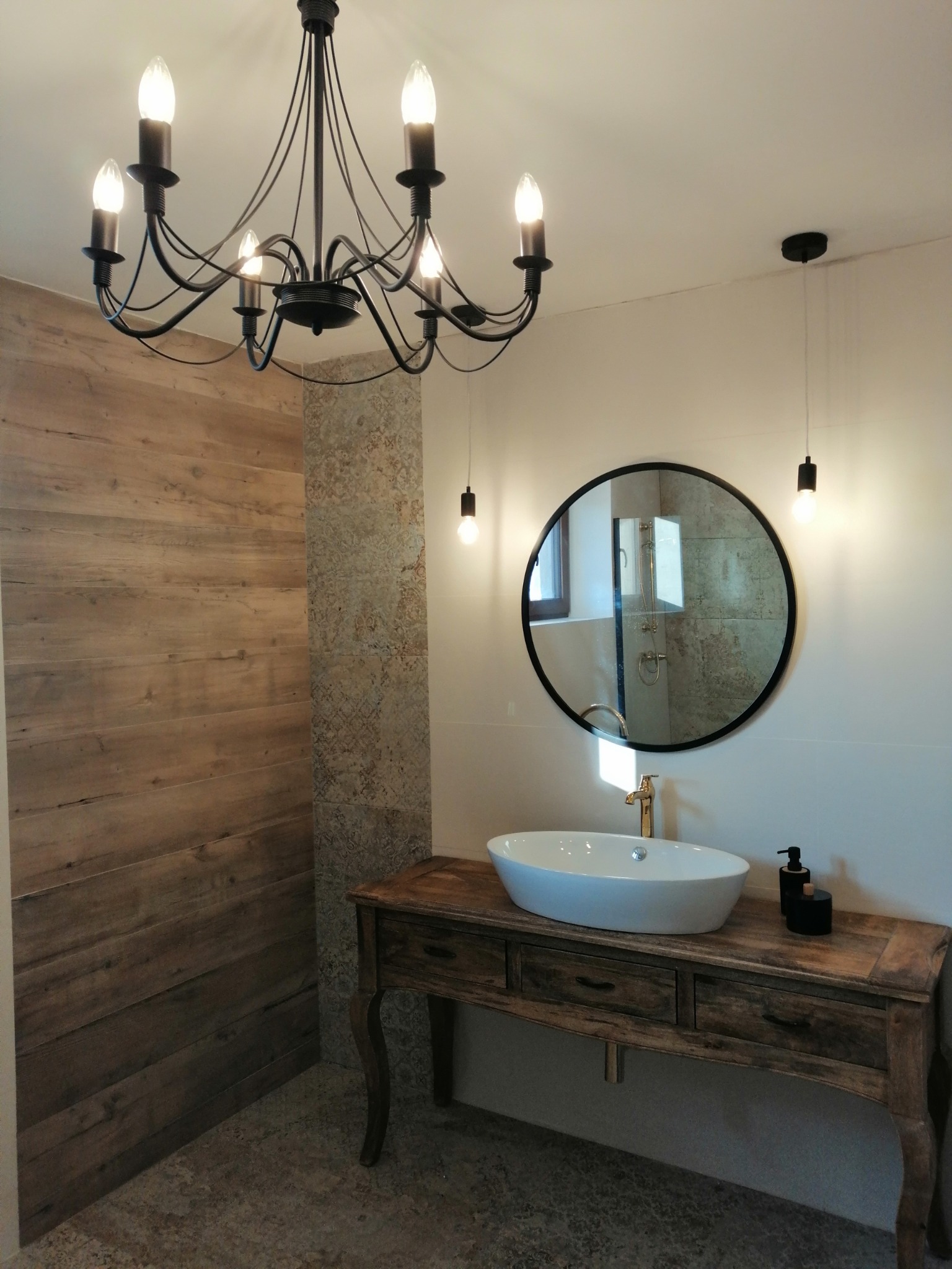 Nowoczesna łazienka z drewnianą ścianą, umywalką nablatową na postarzanej szafce z szufladami, okrągłym lustrem i czarnym żyrandolem