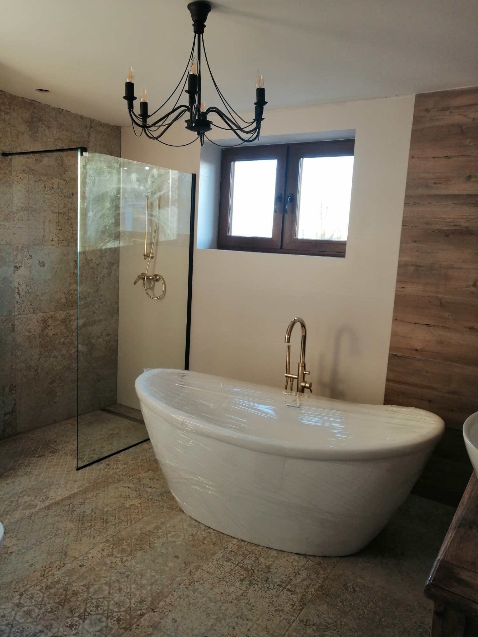 Nowoczesna łazienka z wolnostojącą białą wanną owiniętą folią ochronną, złotą armaturą i kabiną prysznicową typu walk-in z brązowymi płytkami imitującymi kamień, oświetlona czarnym, ozdobnym...