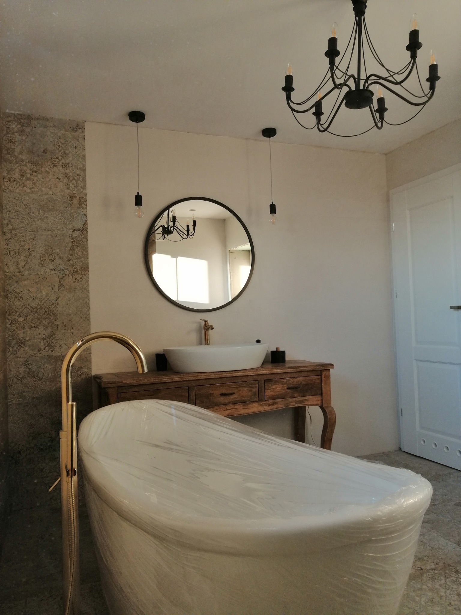 Nowa łazienka z wolnostojącą wanną owiniętą folią, drewnianą szafką pod umywalką i ozdobnym żyrandolem, płytki imitujące wzorzysty kamień na ścianie.